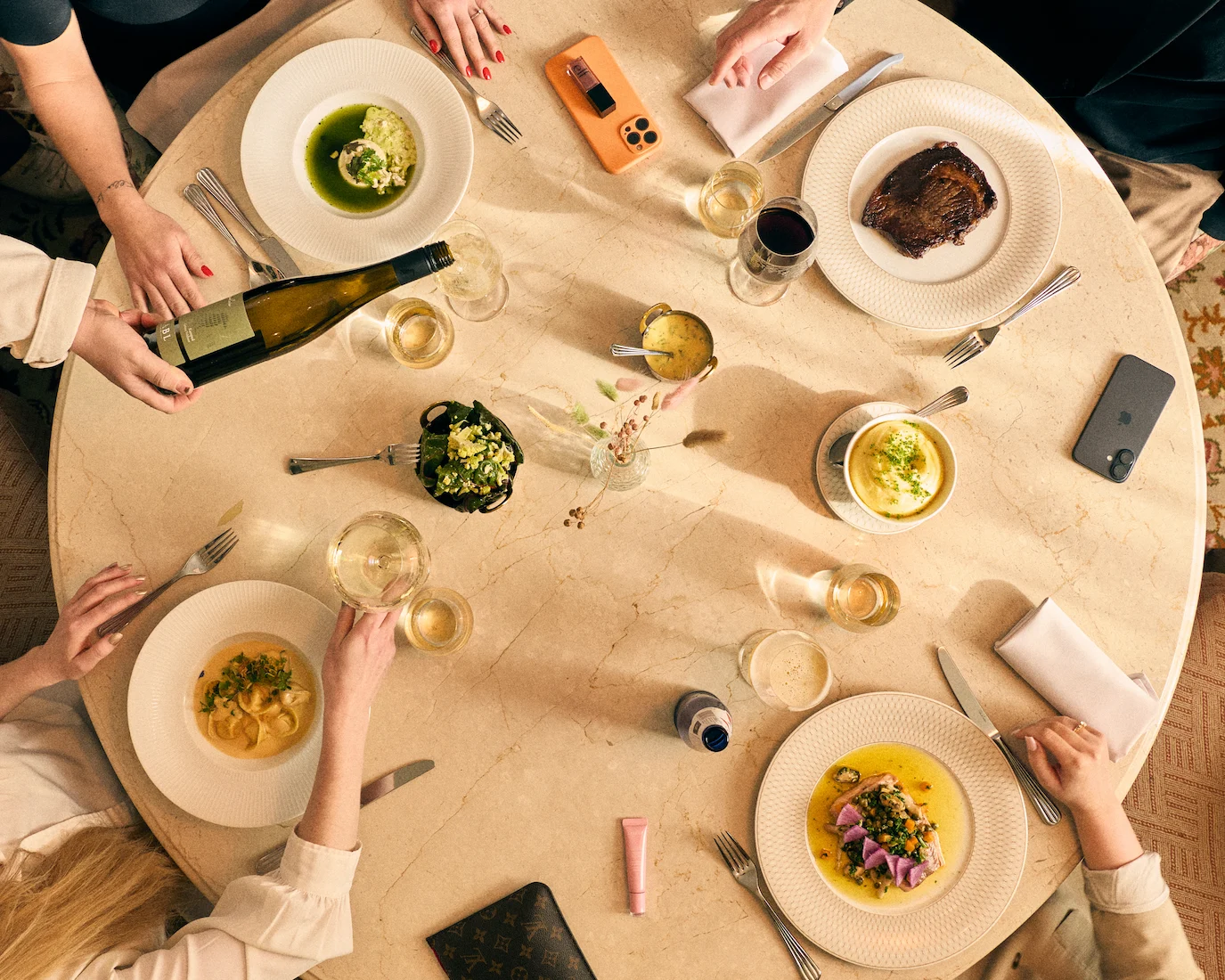 Flera personer äter middag runt ett runt bord med olika rätter, vin och mobiltelefoner