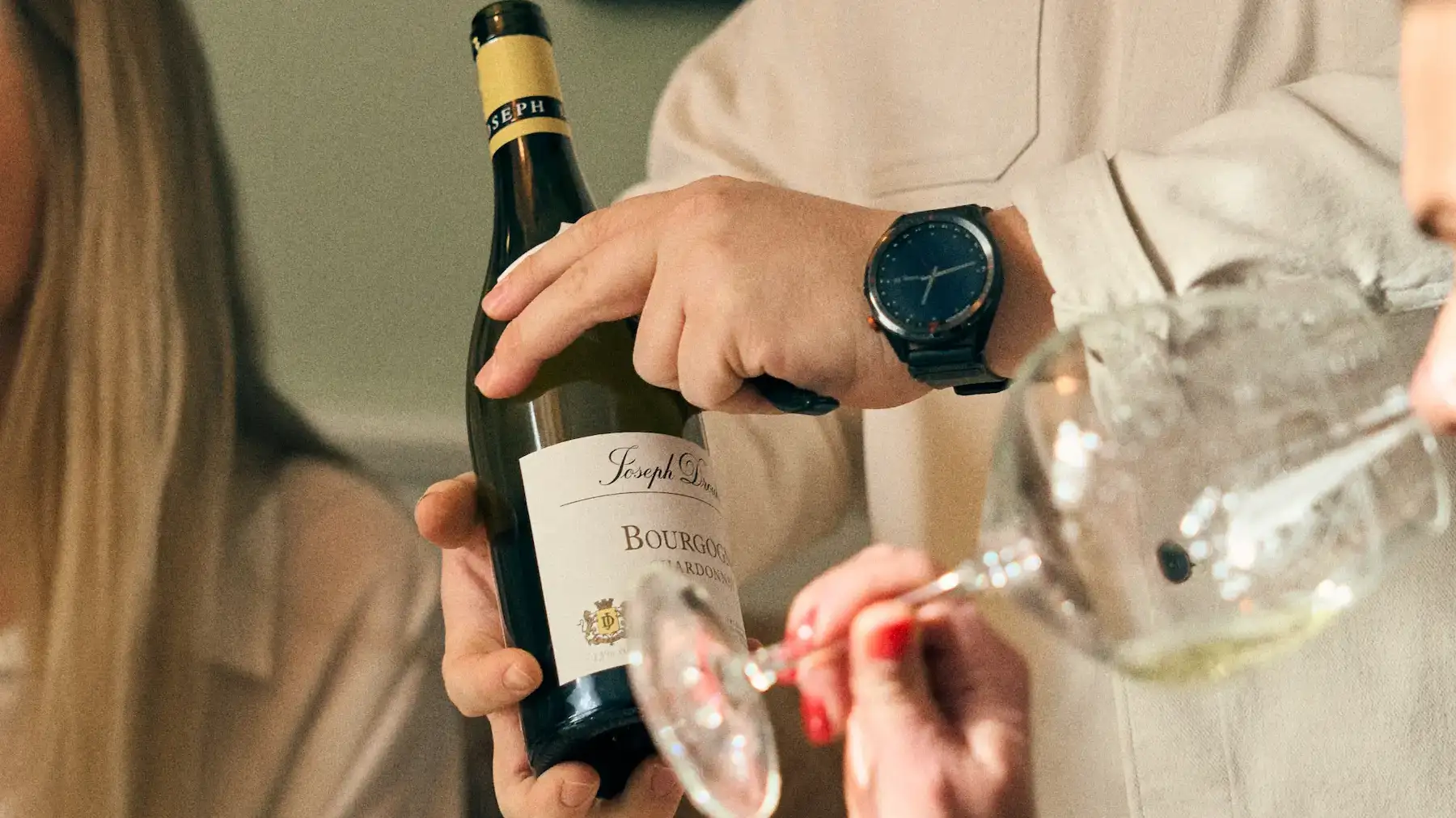 Person håller upp en flaska Bourgogne Chardonnay och häller upp vin i glas under en middag