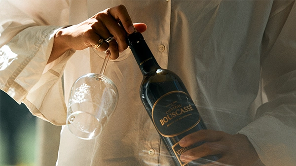 Photo of a person in a cream shirt holding a wine glass in one hand and a bottle of Château Bouscassé wine in the other, set against a warm, softly lit background.